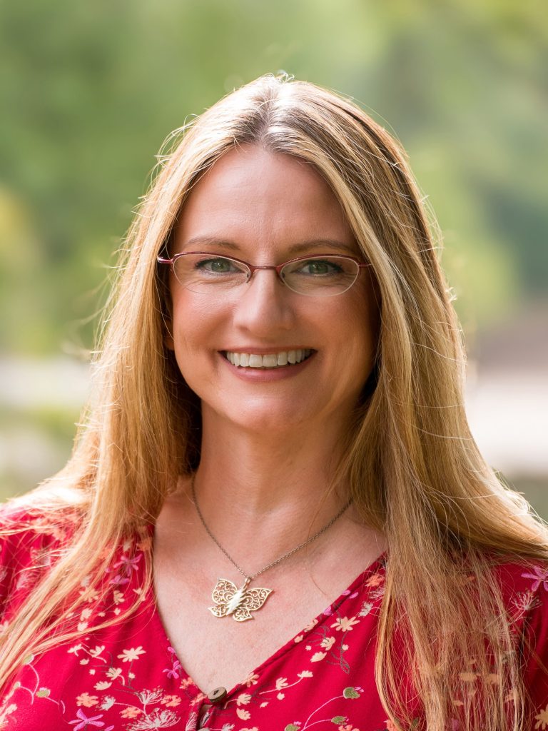 Smiling woman with glasses and long hair, wearing a floral red blouse and butterfly necklace, representing a neurodivergent therapist promoting empowerment for ADHD and Autism.